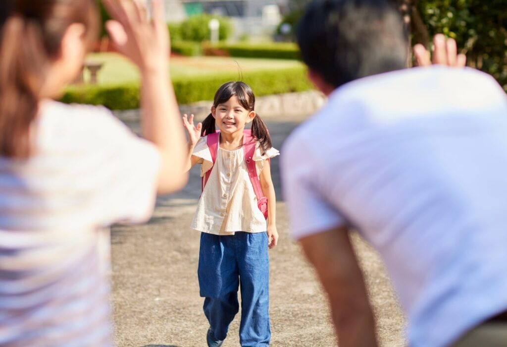 ランドセルを背負った女の子が、迎えに来た保護者に笑顔で手をふっている場面