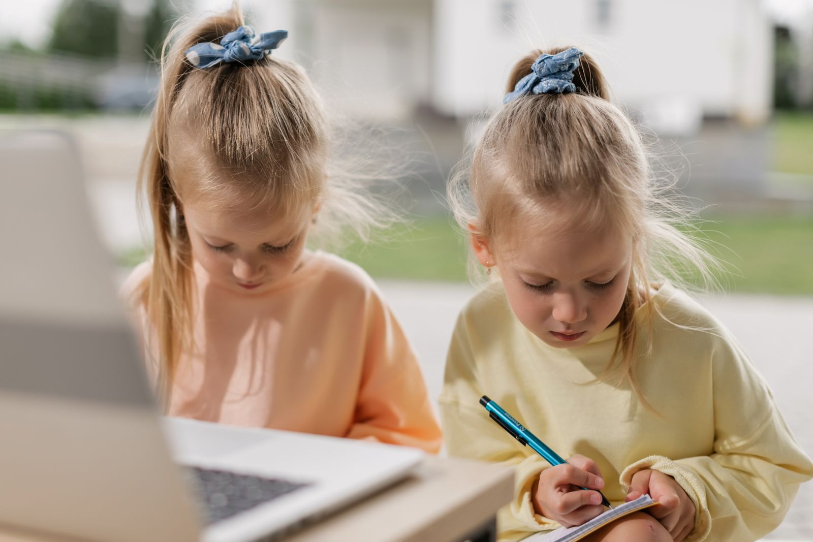 Two children sitting side by side outdoors with a laptop and notebooks
