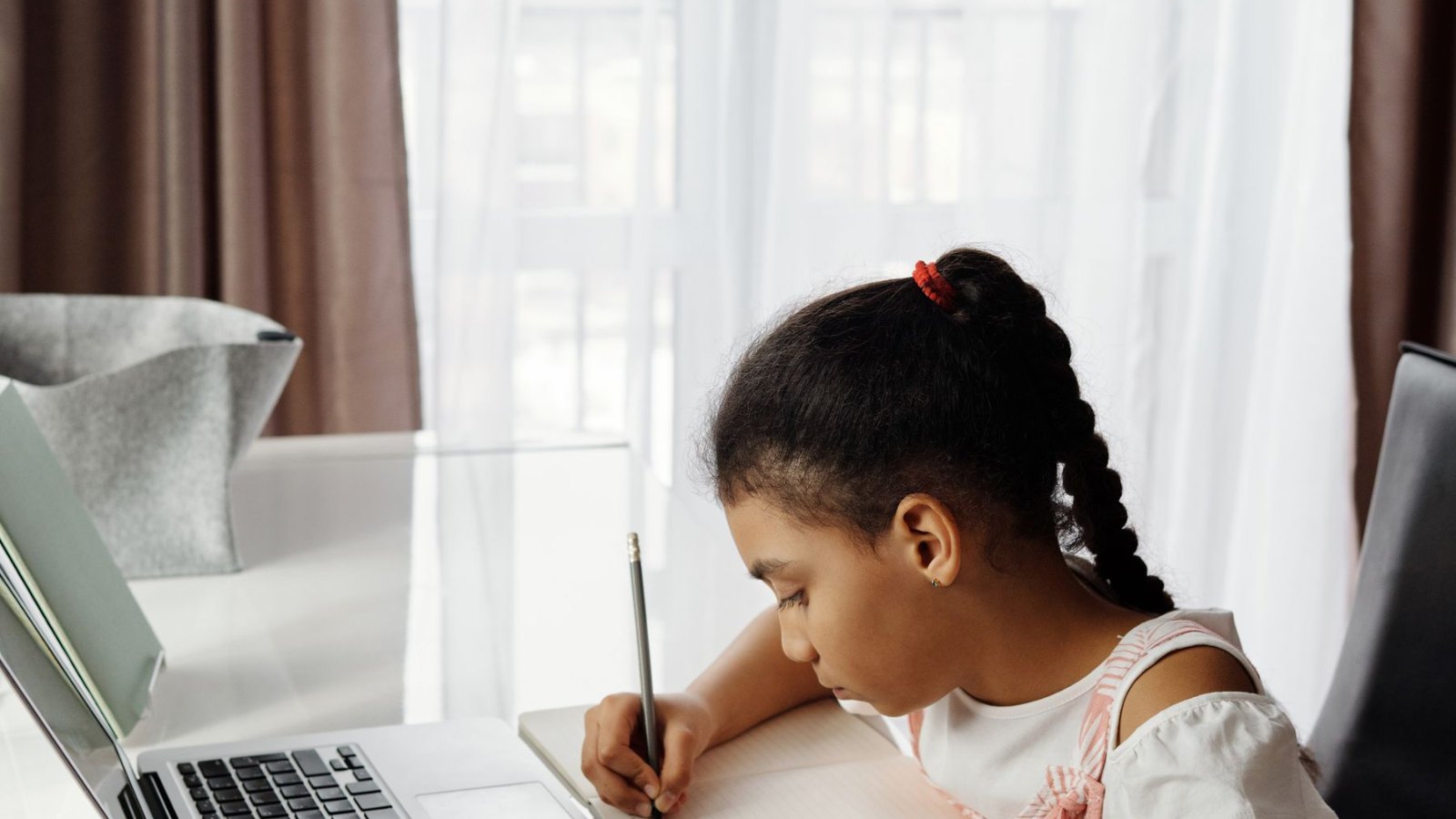 明るい部屋で、ノートパソコンのそばに座って書きものをする子ども