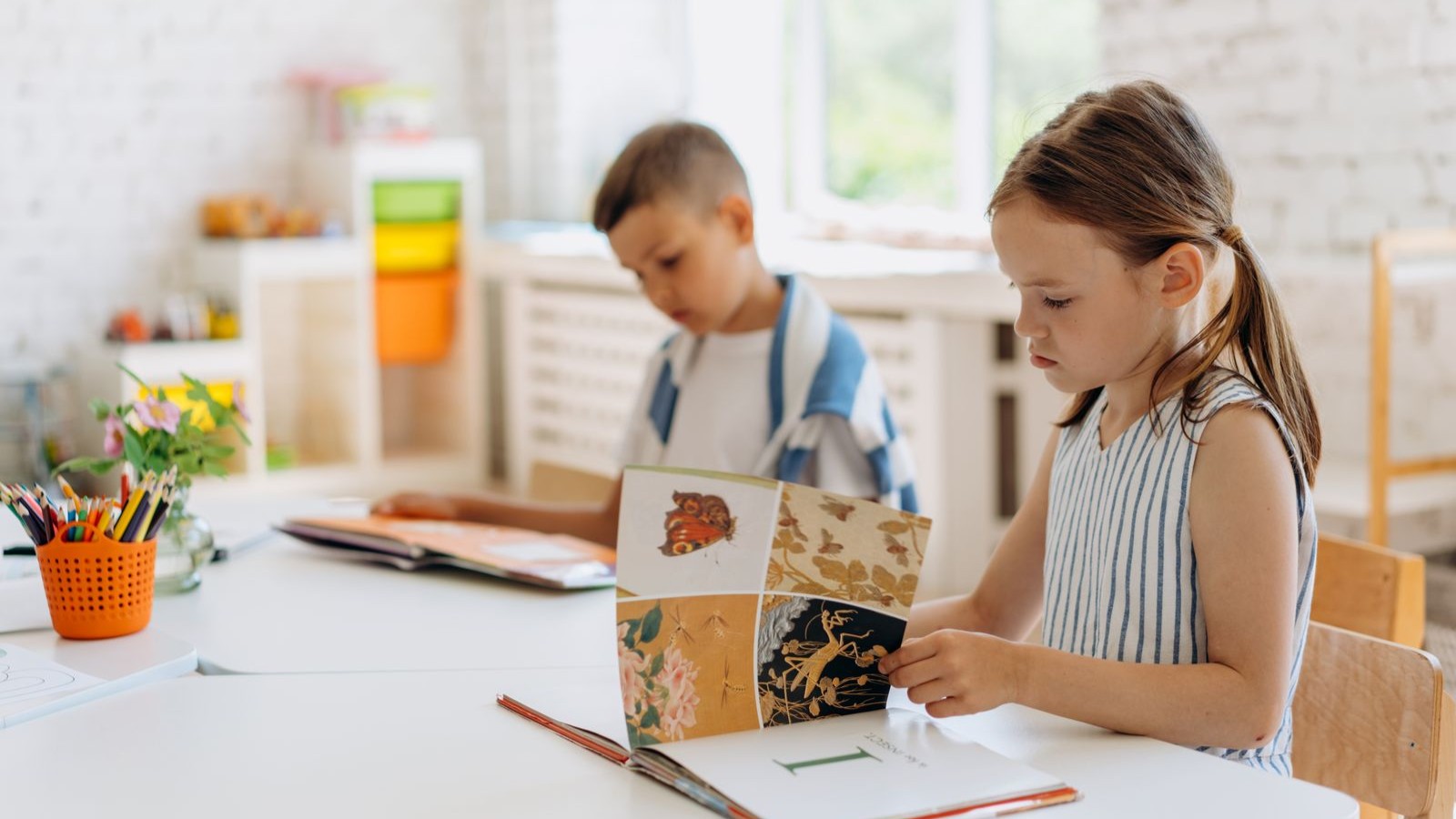 Two children sitting at a bright table looking at an open picture book together
