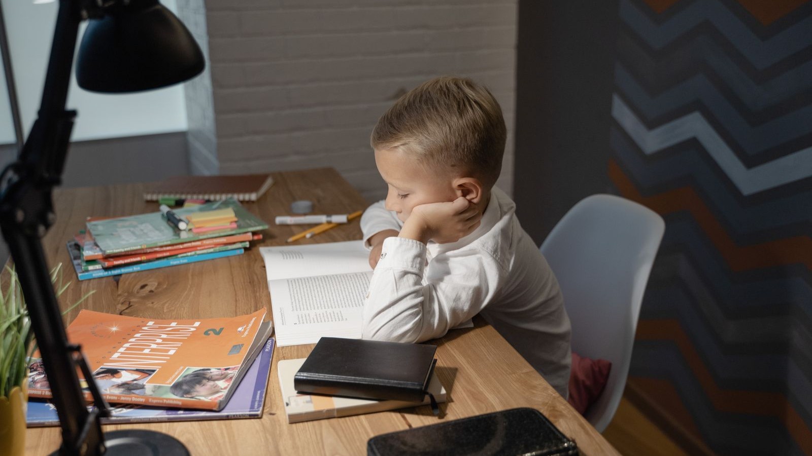 A boy reading a book at a desk by lamplight with study materials